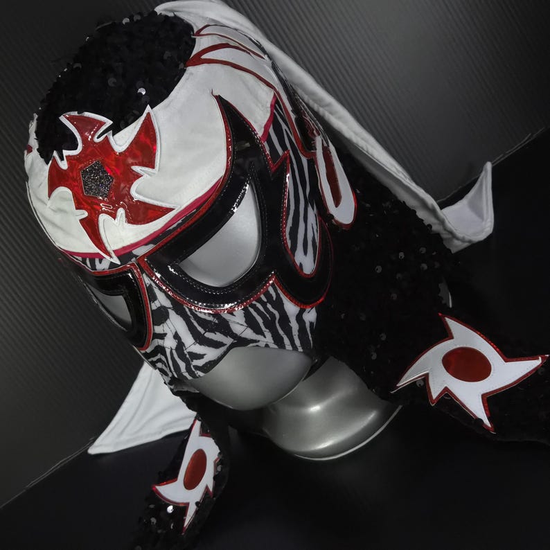 Puede incluir: Una m&aacute;scara de lucha libre con un dise&ntilde;o llamativo. La m&aacute;scara presenta una base blanca con detalles rojos y negros, incluido un estampado de cebra en la cara. Est&aacute; adornada con lentejuelas y una capucha blanca.