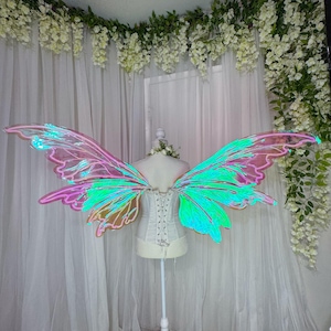 May include: Large iridescent fairy wings with a translucent, shimmering surface that shifts between pink, green, and blue hues. The wings feature a defined, structured frame and are attached to a white corset worn on a mannequin stand.