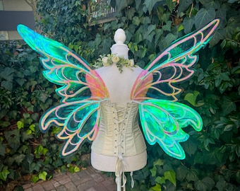 May include: A mannequin displays iridescent fairy wings with a cream-colored corset. The wings have a vibrant color gradient of green, blue, and pink. The corset is laced up in the back and adorned with a floral arrangement at the neckline.