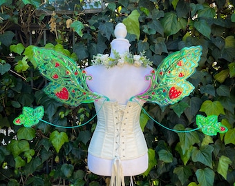 May include: A white corset with a lace-up back, adorned with large, iridescent green fairy wings featuring red strawberry patterns and intricate filigree details. Two smaller matching leaf-shaped wing accents extend outward, and a floral crown sits atop the mannequin.