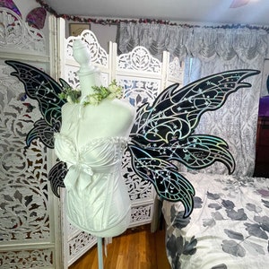 May include: A white corset with a bow and decorative black fairy wings with a celestial design. The wings are attached to a mannequin. The wings have a shimmering, iridescent finish and are decorated with stars and crescent moons.