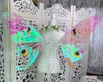 May include: A pair of iridescent butterfly wings with green, pink, and yellow accents. The wings are attached to a white corset and are displayed on a white mannequin.