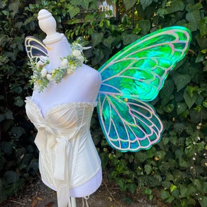 May include: A white satin corset with a bow and lace trim, paired with a green iridescent butterfly fairy wings costume.