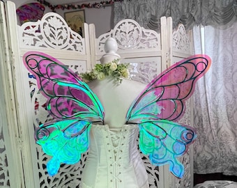 May include: A white corset with a lace-up front is displayed with iridescent butterfly wings. The wings have a gradient of pink, green, and blue hues. The wings are attached to a mannequin, set against a decorative white screen.