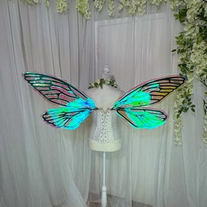 May include: Iridescent fairy wings in green and blue hues are displayed on a white dress form. The wings feature a delicate, veined pattern and are set against a backdrop of white curtains and floral decorations.