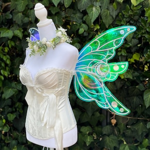 May include: A white satin corset with a bow and a white flower crown. The corset is worn on a mannequin with iridescent green fairy wings.