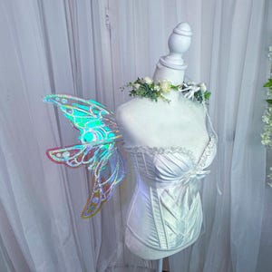 May include: A white satin corset with a sweetheart neckline and decorative boning. Iridescent fairy wings with a glittery finish are attached to the mannequin. A floral garland adorns the neck.