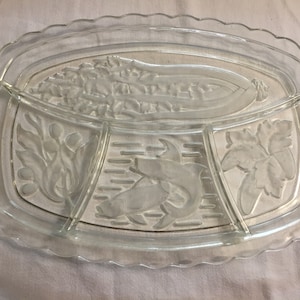 May include: Clear glass serving platter with three divided sections and a scalloped edge. The sections feature embossed designs of vegetables and fish. A decorative piece for serving appetizers or snacks.