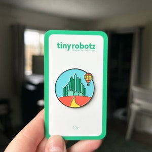 Könnte beinhalten: Eine grüne und gelbe Emaille-Anstecknadel mit der Smaragdstadt aus dem Zauberer von Oz. Die Anstecknadel befindet sich auf einer weißen Karte mit dem Text "tinyrobotz designed by candy burger" und "Oz" unten.