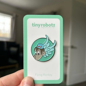 May include: A green and blue enamel pin featuring a cartoon monkey with wings, wearing a blue hat with a red stripe. The pin is on a white card with the text "tinyrobotz designed by candy burger" and "Flying Monkey".