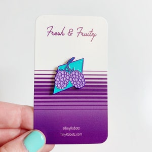 May include: A purple and teal enamel pin featuring two blackberries with a "Fresh & Fruity" text on the card. The card has a purple and white striped background with the text "@TinyRobotz TinyRobotz.com" at the bottom.