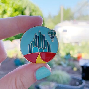 Könnte beinhalten: Eine runde Emaille-Anstecknadel mit einer Darstellung der Smaragdstadt aus dem Zauberer von Oz. Die Anstecknadel ist blau, grün, rot und gelb. Ein Heißluftballon fliegt über der Stadt.