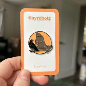 May include: A black crow perched on a brown witch hat with a yellow and red patch. The hat is on a yellow background with the text "tiny robotz designed by cindy burger" and "Scarecrow" below the image.