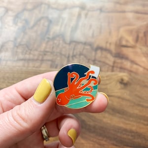 May include: A gold-toned enamel pin featuring an orange octopus with white eyes, holding a white flag, on a blue and green background.