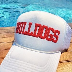 May include: White trucker hat with a red "BULLDOGS" logo on the front. The hat is made of a mesh material and has a white adjustable strap.