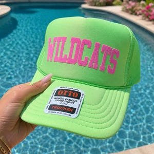 May include: A neon green trucker hat with the word "WILDCATS" in pink block letters. The hat has a mesh back and a curved brim. A white tag with black text is attached to the brim.