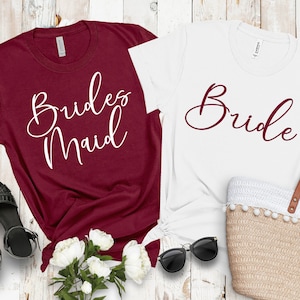May include: Two t-shirts, one burgundy and one white, with text. The burgundy shirt says "Brides Maid" in white script. The white shirt says "Bride" in burgundy script. Accessories include black sunglasses, black sandals, and a straw tote bag.