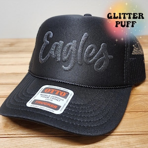 May include: Black trucker hat with a black mesh back and a silver glitter puff embroidered "Eagles" on the front. The hat has a snap closure and a label that reads "OTTO World Famous Foam Front OSFM Trucker Durable Snap Closure".