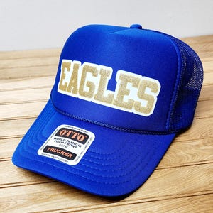 May include: A royal blue trucker hat with a gold and white "EAGLES" logo on the front. The hat has a mesh back and a snap closure.