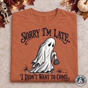 Könnte beinhalten: Ein rostorangefarbenes T-Shirt mit einer schwarzen Grafik eines Geistes, der eine kleine Handtasche trägt. Der Text über dem Geist lautet SORRY I'M LATE. und der Text darunter lautet I DIDN'T WANT TO COME. Das Design ist in schwarzer Tinte gehalten.