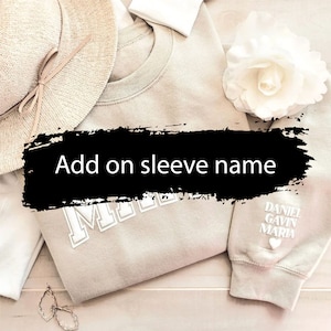 May include: A light beige sweatshirt with the word "Mom" embroidered on the front. The sweatshirt has a white heart and the names "Daniel", "Gavin", and "Maria" embroidered on the sleeve. The sweatshirt is laid out on a light wood surface with a white flower and a straw hat.