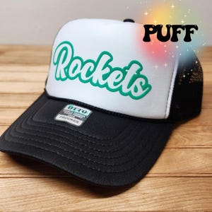 May include: A white and black trucker hat with a green "Rockets" logo on the front. The hat has a black mesh back and a black bill. The hat has a label that says "OTTO World Famous Foam Front Trucker".
