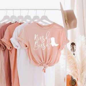 Op de afbeelding: Een roze T-shirt met de tekst "Nash Bash" in witte cursieve letters en een wit silhouet van een cowboylaars. Het shirt hangt aan een witte hanger.