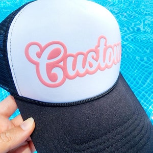 以下が含まれることがあります： 前面にピンクの「Custom」ロゴが付いた白黒のトラッカーハットです。