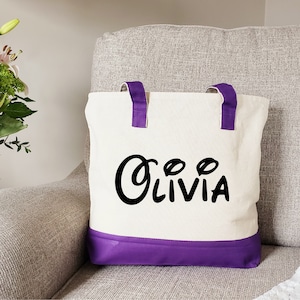 May include: A white canvas tote bag with purple handles and a purple bottom. The bag has the name "Olivia" written in black cursive letters.