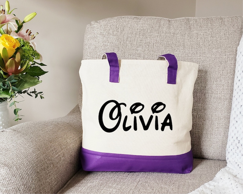 May include: A white canvas tote bag with purple handles and a purple bottom. The bag has the name "Olivia" written in black cursive letters.