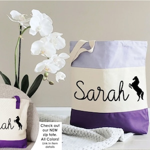 K&ouml;nnte beinhalten: Zwei Canvas-Tragetaschen mit lilafarbener Einfassung. Die gr&ouml;&szlig;ere Tasche tr&auml;gt den Namen "Sarah" und eine schwarze Silhouette eines Pferdes. Die kleinere Tasche hat das gleiche Design, ist aber kleiner. Die Taschen befinden sich auf einer wei&szlig;en Oberfl&auml;che mit einer wei&szlig;en Orchidee im Hintergrund.