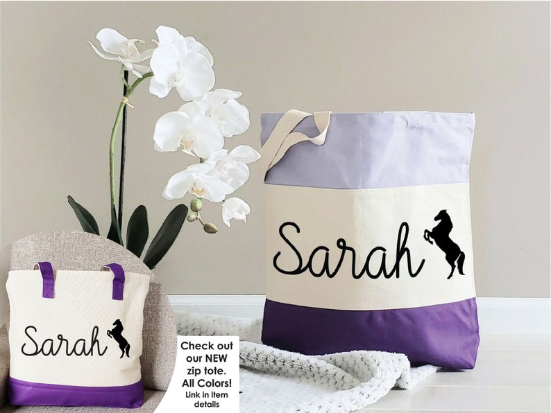 K&ouml;nnte beinhalten: Zwei Canvas-Tragetaschen mit lilafarbener Einfassung. Die gr&ouml;&szlig;ere Tasche tr&auml;gt den Namen "Sarah" und eine schwarze Silhouette eines Pferdes. Die kleinere Tasche hat das gleiche Design, ist aber kleiner. Die Taschen befinden sich auf einer wei&szlig;en Oberfl&auml;che mit einer wei&szlig;en Orchidee im Hintergrund.