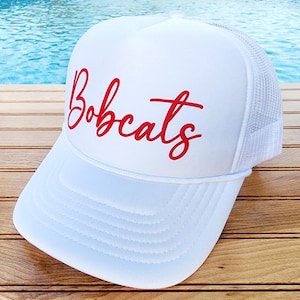 以下が含まれることがあります： 赤い刺繍の「Bobcats」ロゴが付いた白いトラッカーハット。帽子はメッシュバックと白いフロントです。