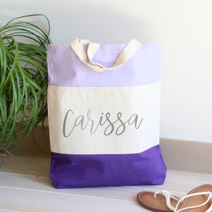 K&ouml;nnte beinhalten: Eine lila, wei&szlig;e und lavendelfarbene Canvas-Tragetasche mit dem Namen "Carissa" in Silber auf der Vorderseite.