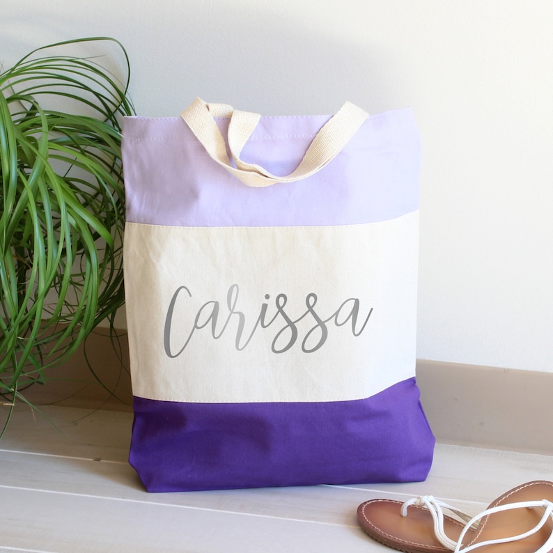 K&ouml;nnte beinhalten: Eine lila, wei&szlig;e und lavendelfarbene Canvas-Tragetasche mit dem Namen "Carissa" in Silber auf der Vorderseite.