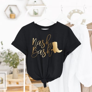Op de afbeelding: Zwart T-shirt met gouden lettertype dat "Nash Bash" zegt en een gouden silhouet van een cowboylaars.