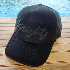 以下が含まれることがあります： 黒のメッシュバックと「Knights」というエンボス加工された黒い刺繍ロゴが付いた、黒いトラッカーハットです。