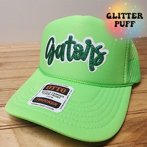May include: A bright green trucker hat with the word "Gators" in a glittery green script. The hat has a mesh back and a curved brim. The hat also has a tag that says "OTTO WORLD FAMOUS FOAM FRONT TRUCKER". The text "GLITTER PUFF" is in the upper right corner.