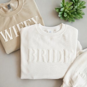 May include: Two white sweatshirts with the words "WIFFY" and "BRIDE" in raised white lettering. The sweatshirts are folded and laid out on a light gray surface.