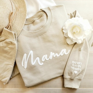 May include: A beige sweatshirt with the word "Mama" in white script across the front. The sleeve features the names "Daniel, Gavin, Maria" and a heart. A straw hat and a white flower are also visible.