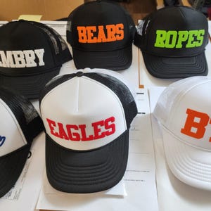 以下が含まれることがあります： メッシュバックパネルが付いた、黒と白の異なる5つのトラッカーハット。ハットには、前面に異なる色の刺繍されたテキストがあります。ハットのテキストは、「Bears」、「Ropes」、「Eagles」、「B」、「Mbrry」です。