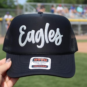 May include: Black trucker hat with the word "Eagles" in white script across the front. The hat has a mesh back and a curved brim. The hat is a foam front style.