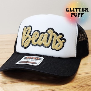 May include: A black and white trucker hat featuring the word Bears in gold glitter script with a black outline on the white foam front panel. The hat includes a black mesh back, a black brim, and a sticker on the brim reading OTTO.