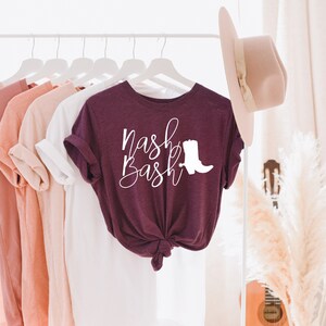 Op de afbeelding: Een maroon t-shirt met de tekst "Nash Bash" in witte cursieve letters en een wit silhouet van een cowboylaars.