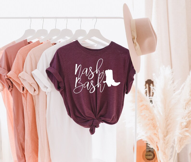Op de afbeelding: Een maroon t-shirt met de tekst "Nash Bash" in witte cursieve letters en een wit silhouet van een cowboylaars.