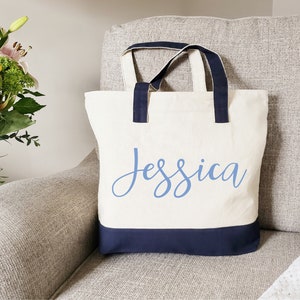 May include: A white canvas tote bag with navy blue handles and trim. The bag has the name "Jessica" written in blue cursive on the front.
