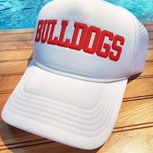 以下が含まれることがあります： 赤い刺繍の「BULLDOGS」ロゴが付いた白いトラッカーハット。帽子は木製の表面の上に置かれています。