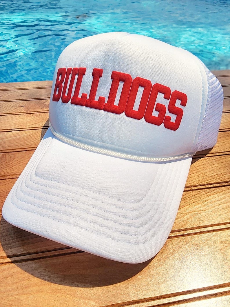 以下が含まれることがあります： 赤い刺繍の「BULLDOGS」ロゴが付いた白いトラッカーハット。帽子は木製の表面の上に置かれています。
