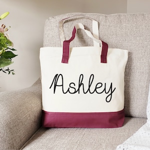 May include: A white canvas tote bag with maroon handles and a maroon bottom border. The bag has the name "Ashley" written in black cursive on the front.