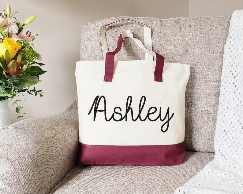 May include: A white canvas tote bag with maroon handles and a maroon bottom border. The bag has the name "Ashley" written in black cursive on the front.
